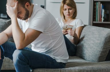 Alt="Upset couple sitting at the sofa. Woman counts last money while her stressed and worried husband holding his head with hands.. Financial family problems concept."